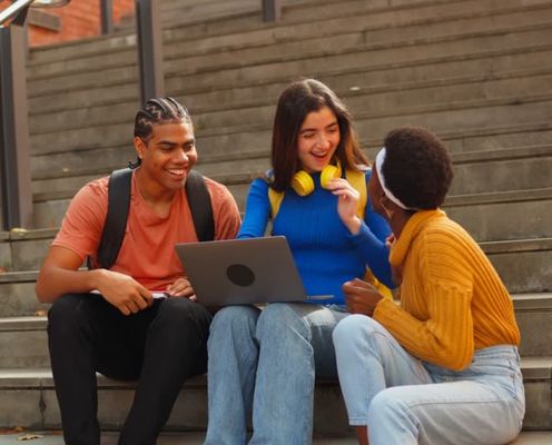 Three young students sitting on steps, smiling and looking at a laptop together